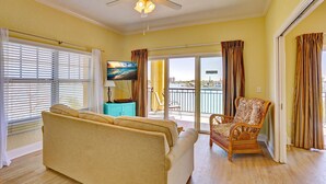 A living room with a sofa, a wicker chair, a painting, and a balcony with a view. - Waterfront Views~ Heated Pool~ Walk to Beach~ Palms of Treasure Island 210 (Treasure Island)