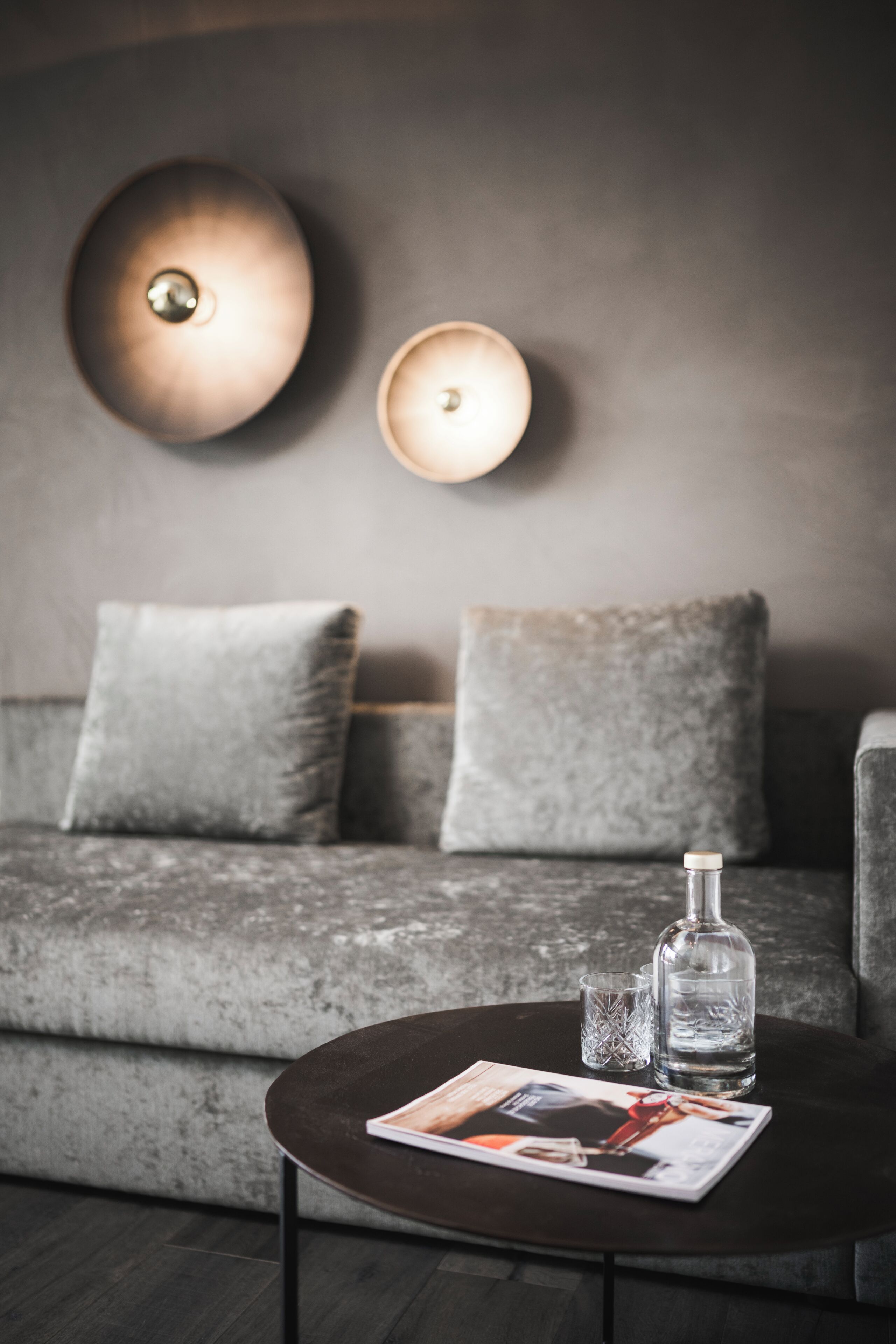 Un salon moderne avec un canapé gris, deux coussins, une table basse ronde sur laquelle se trouvent un magazine, une bouteille et un verre.