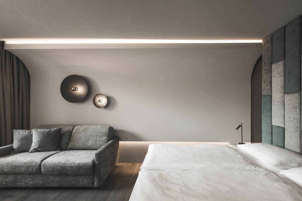 A modern bedroom with a grey sofa, a bed with white linens, and a wall-mounted light fixture.