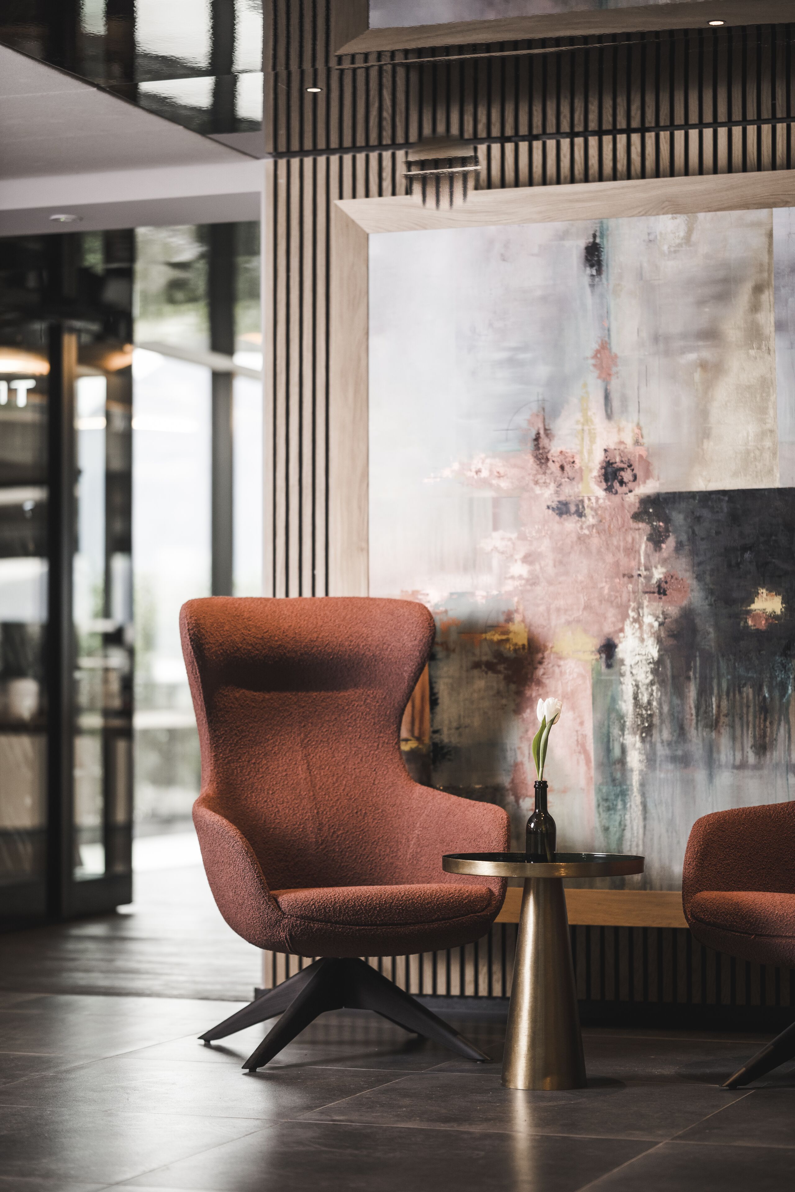 Un interior moderno con un sillón tapizado rojo, una pequeña mesa redonda con un florero y un cuadro abstracto grande en la pared.