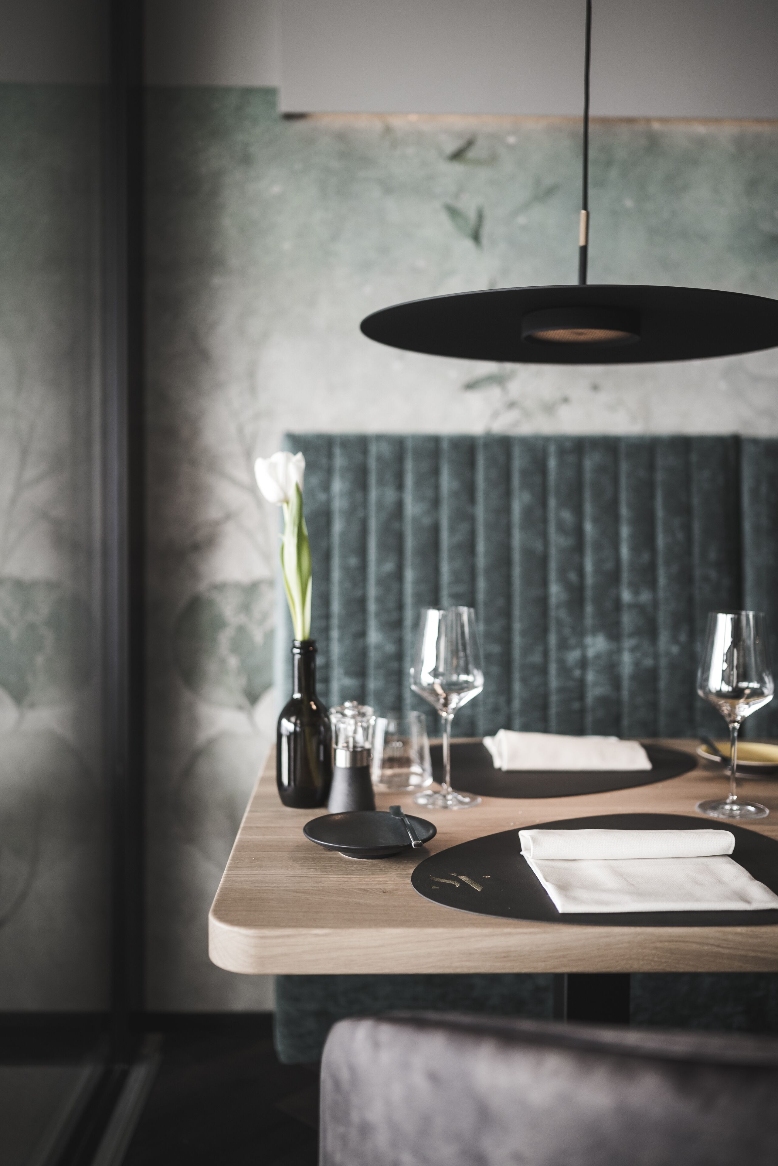 Una mesa de comedor con un posavasos negro, una servilleta blanca, dos copas de vino, una botella y un florero con flores blancas.