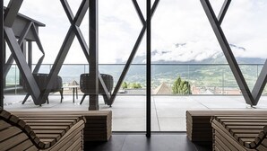 Un balcón con vistas a un paisaje montañoso, bancos de madera y una barandilla de cristal.