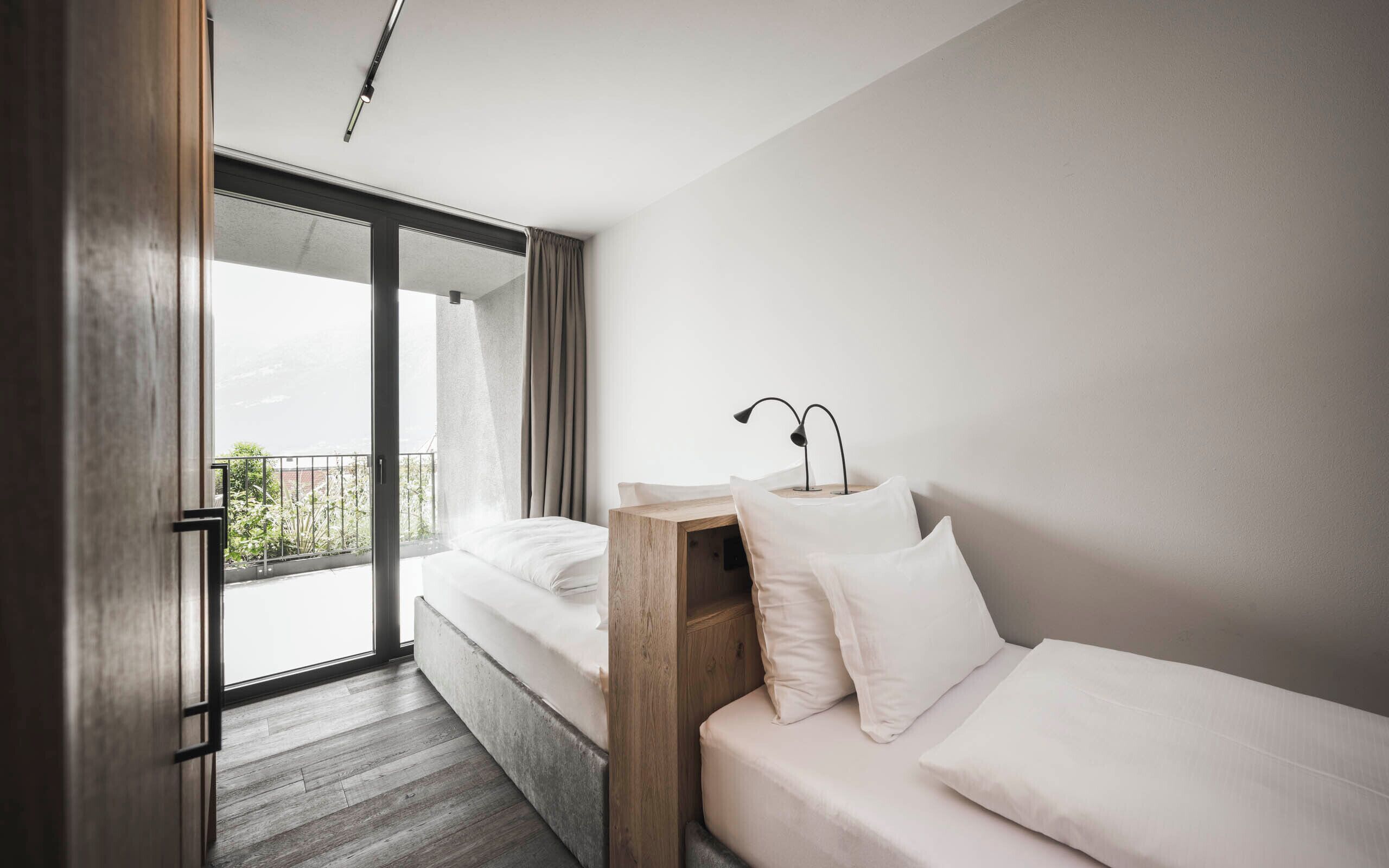 Una habitación de hotel moderna con un amplio ventanal corredero que da a un balcón, una mesita de noche de madera y una cama con ropa blanca.
