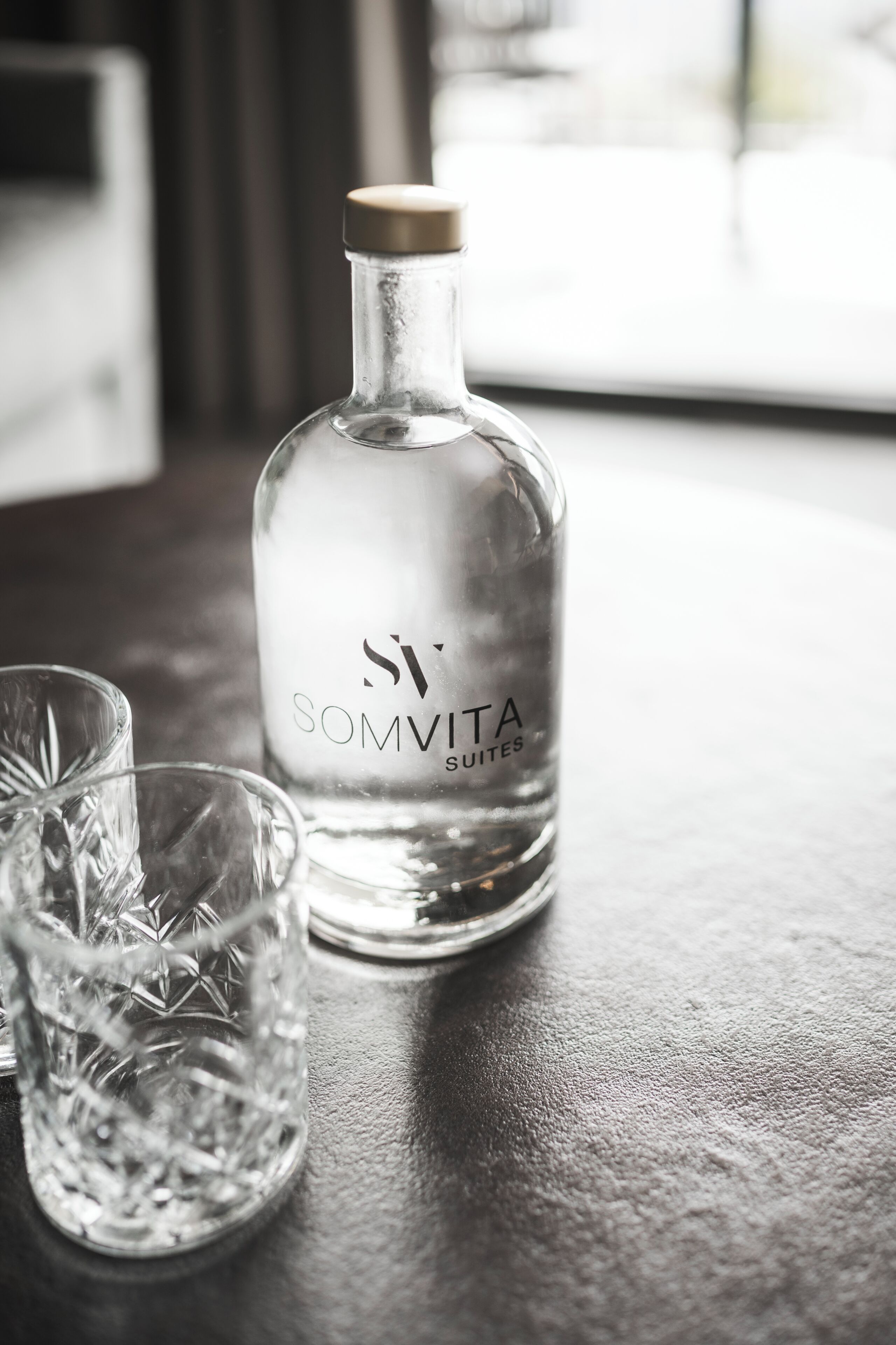 A bottle labeled 'SOMVITA SUITES' and two crystal glasses on a textured surface.
