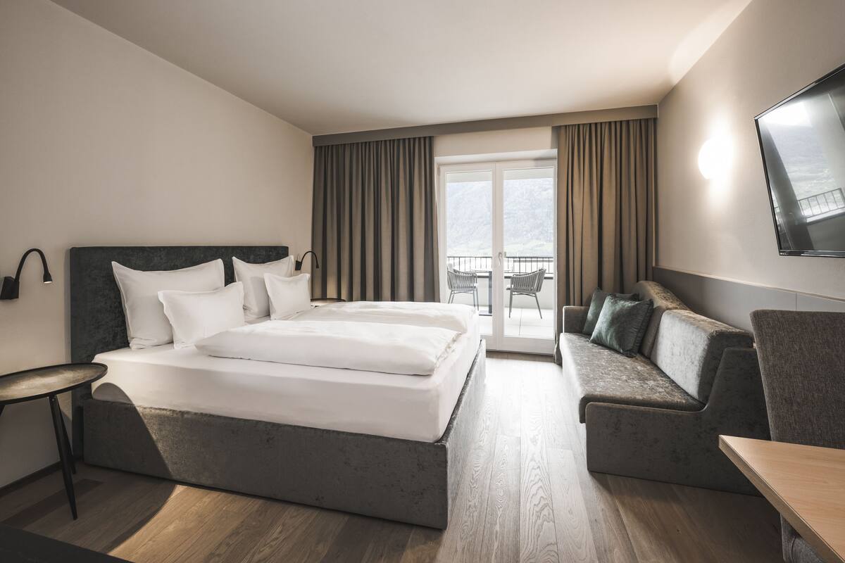 A modern hotel room with a large bed, a sofa, a TV, and a balcony with curtains.
