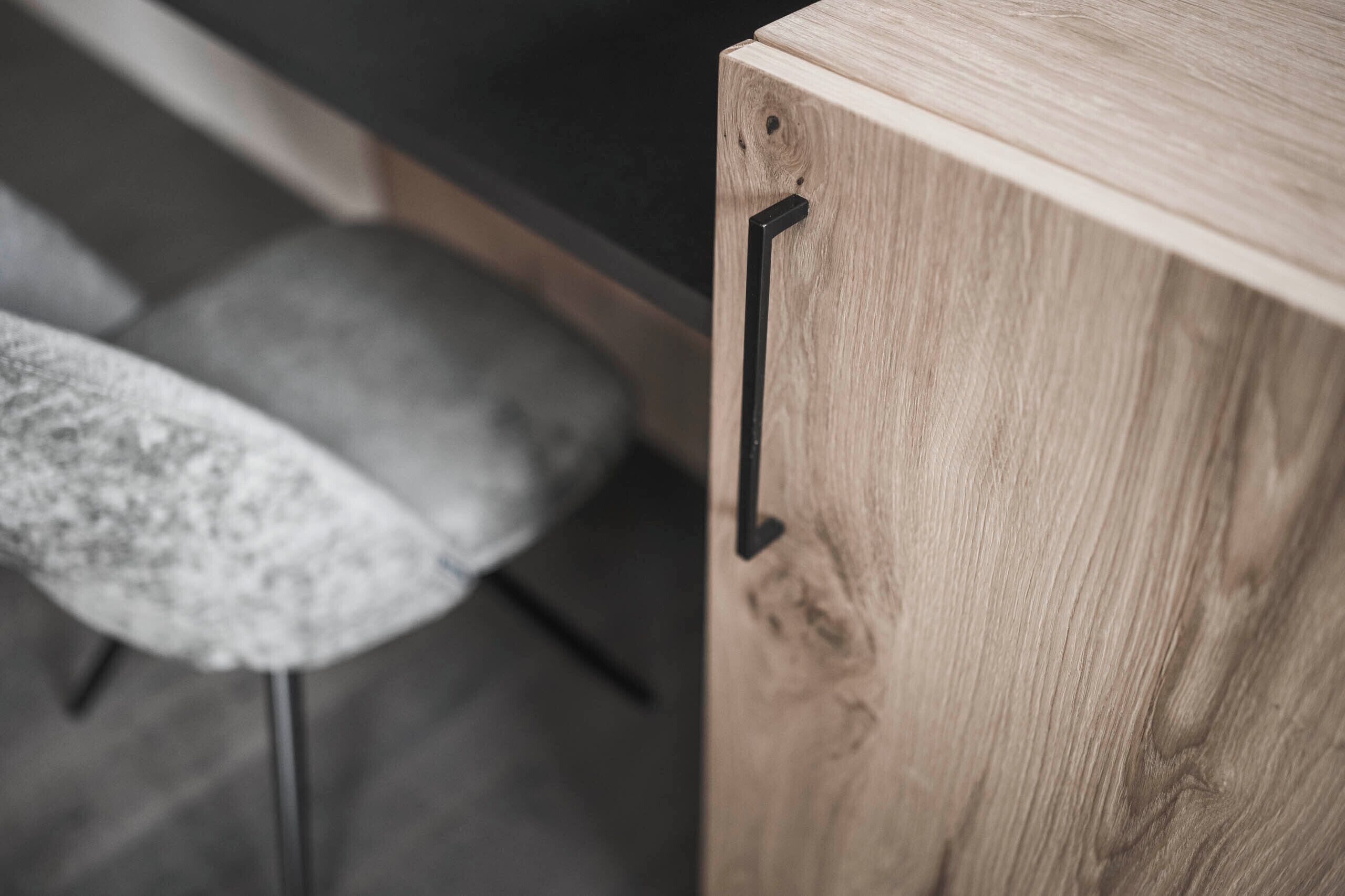 A wooden cabinet with a black handle, a grey cushioned stool, and a dark countertop.