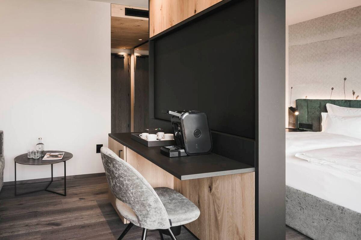 A modern hotel room with a bed, a desk with a coffee machine, a chair, and a small table with glasses and a bottle.