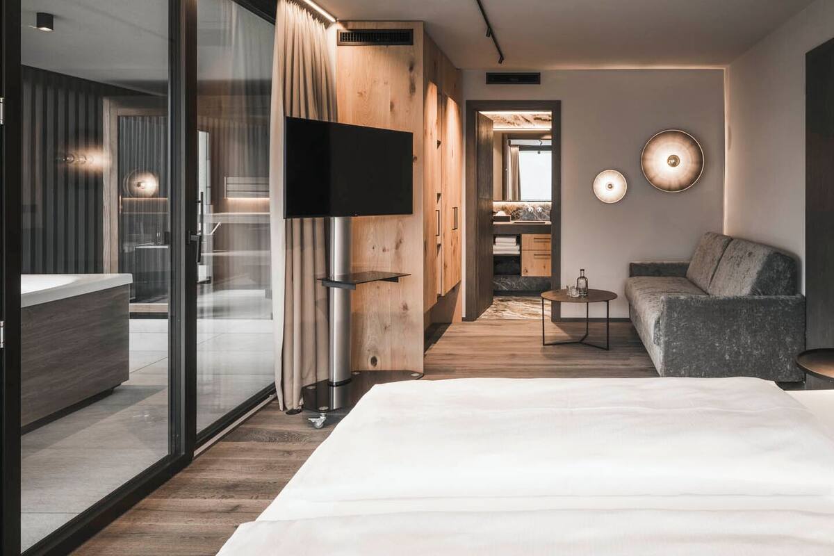 A modern hotel room with a large bed, a seating area with a table, and a view into the kitchen.