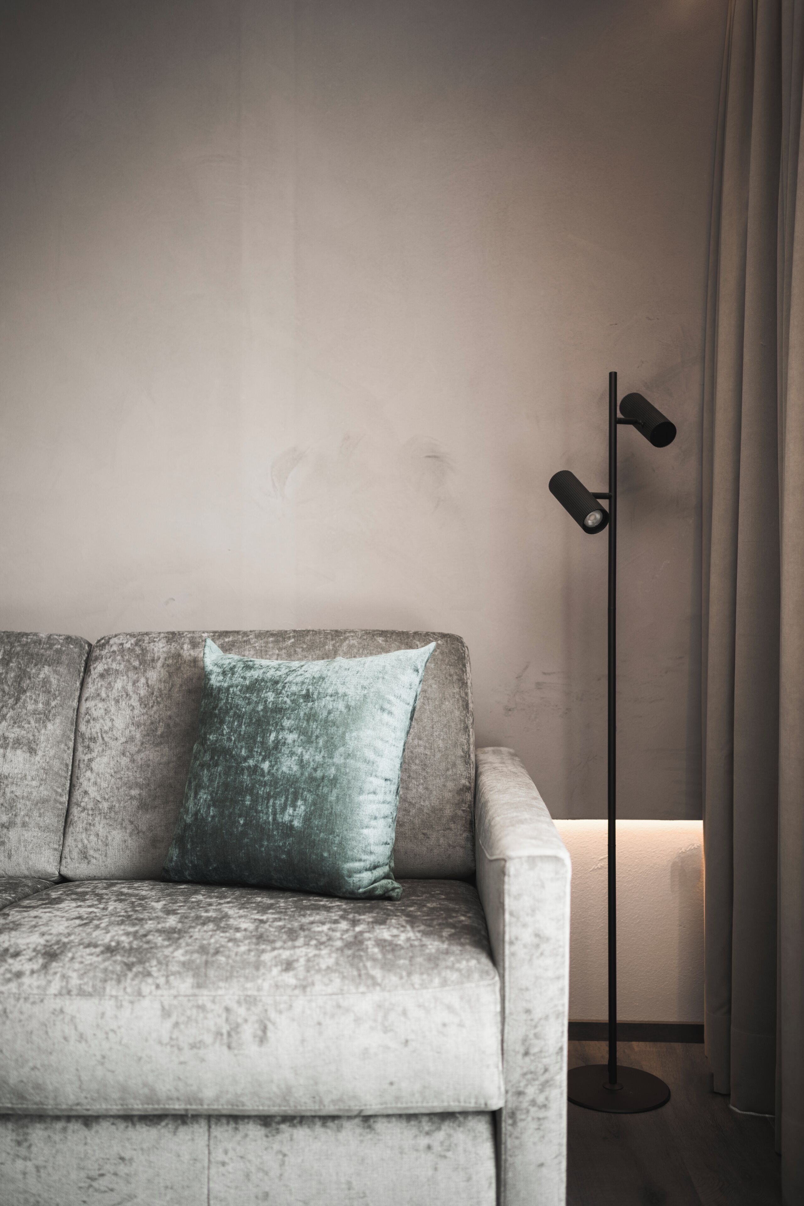 Un canapé recouvert d’un tissu texturé, agrémenté d’un seul coussin, un lampadaire à deux abat-jour et un rideau tiré partiellement sur le côté.