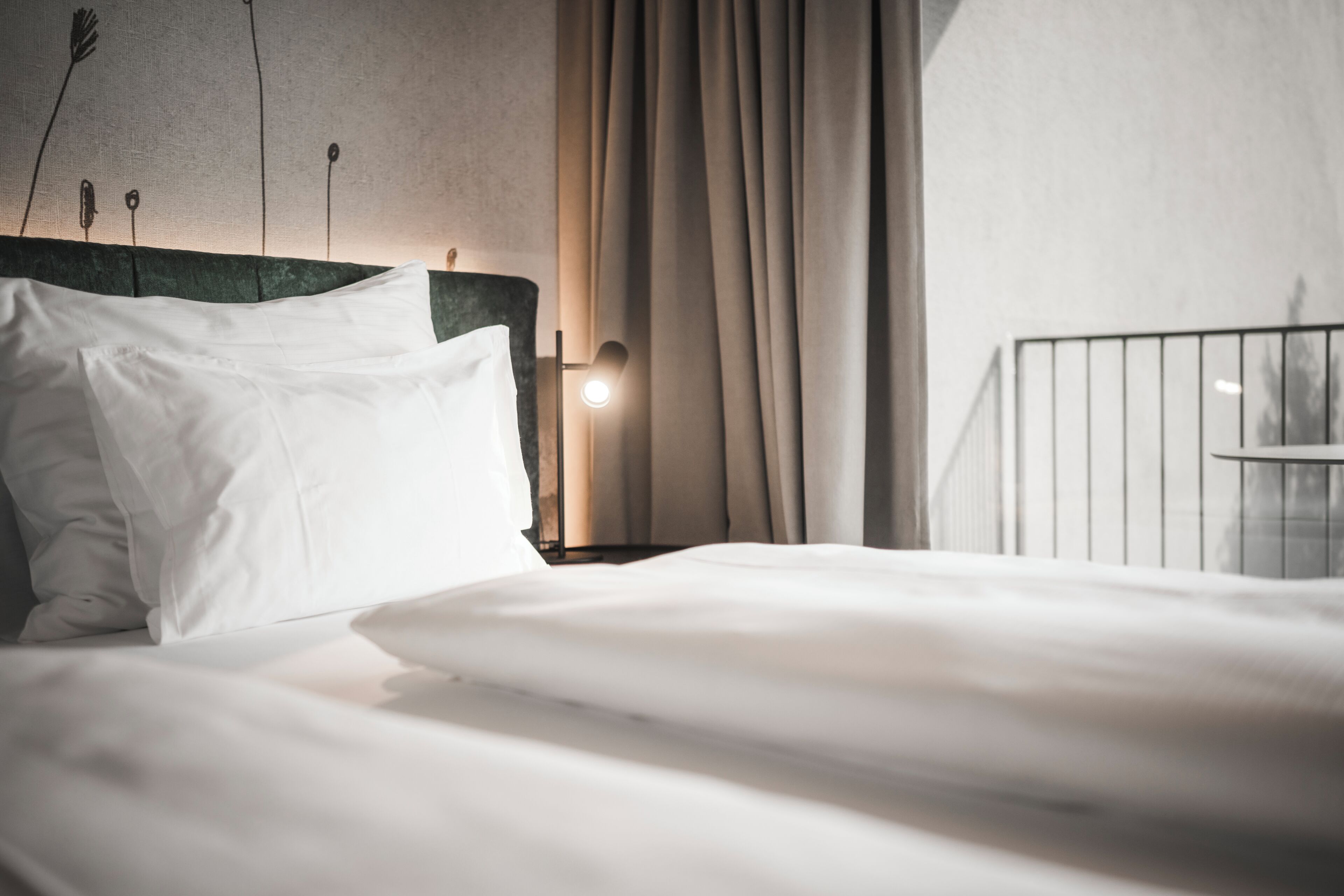 Een net opgemaakt bed met witte lakens, een met groen bekleden hoofdeinde en een bedlampje.