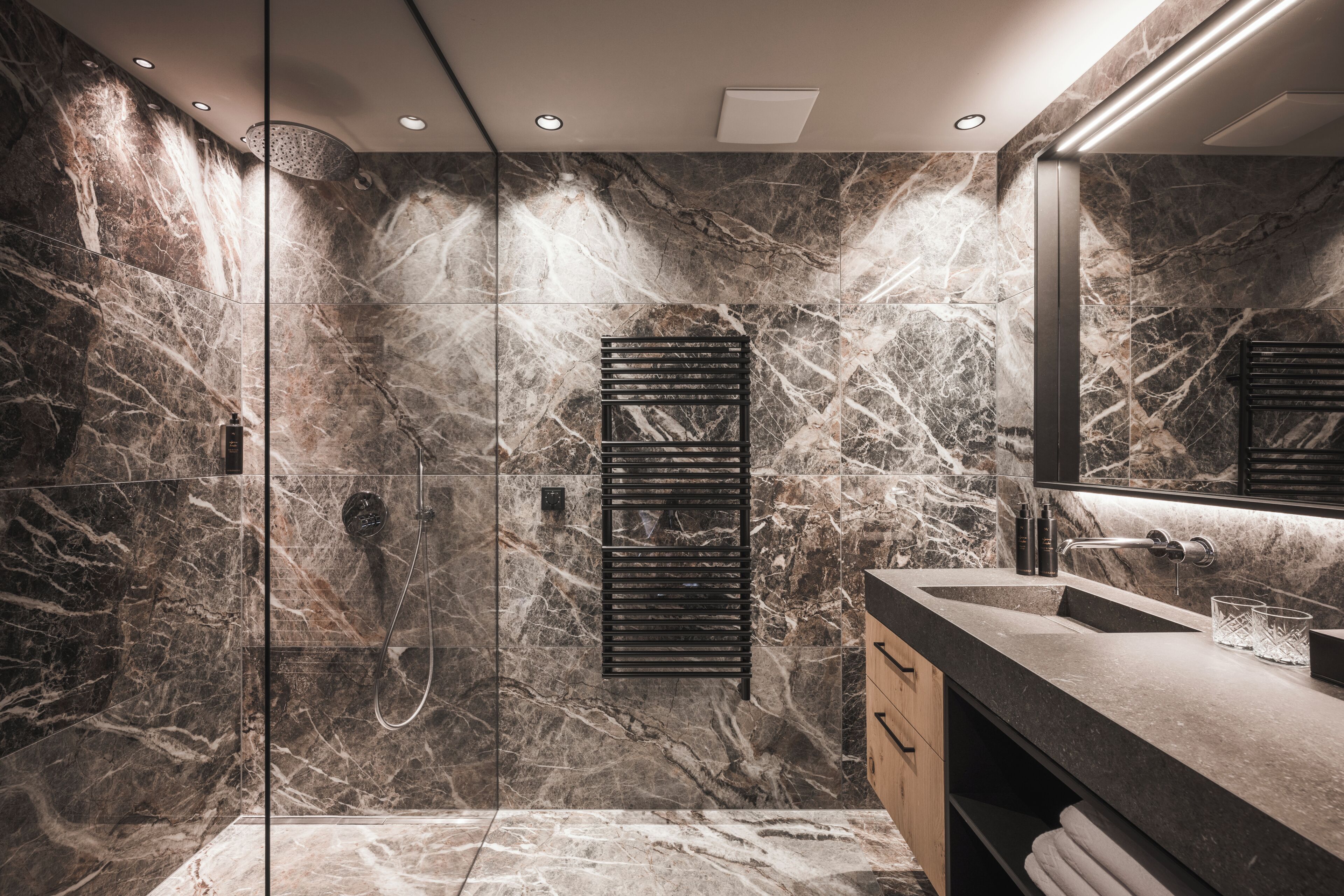 A modern bathroom with a glass-enclosed shower, marble walls, a black towel rack, and a large mirror.