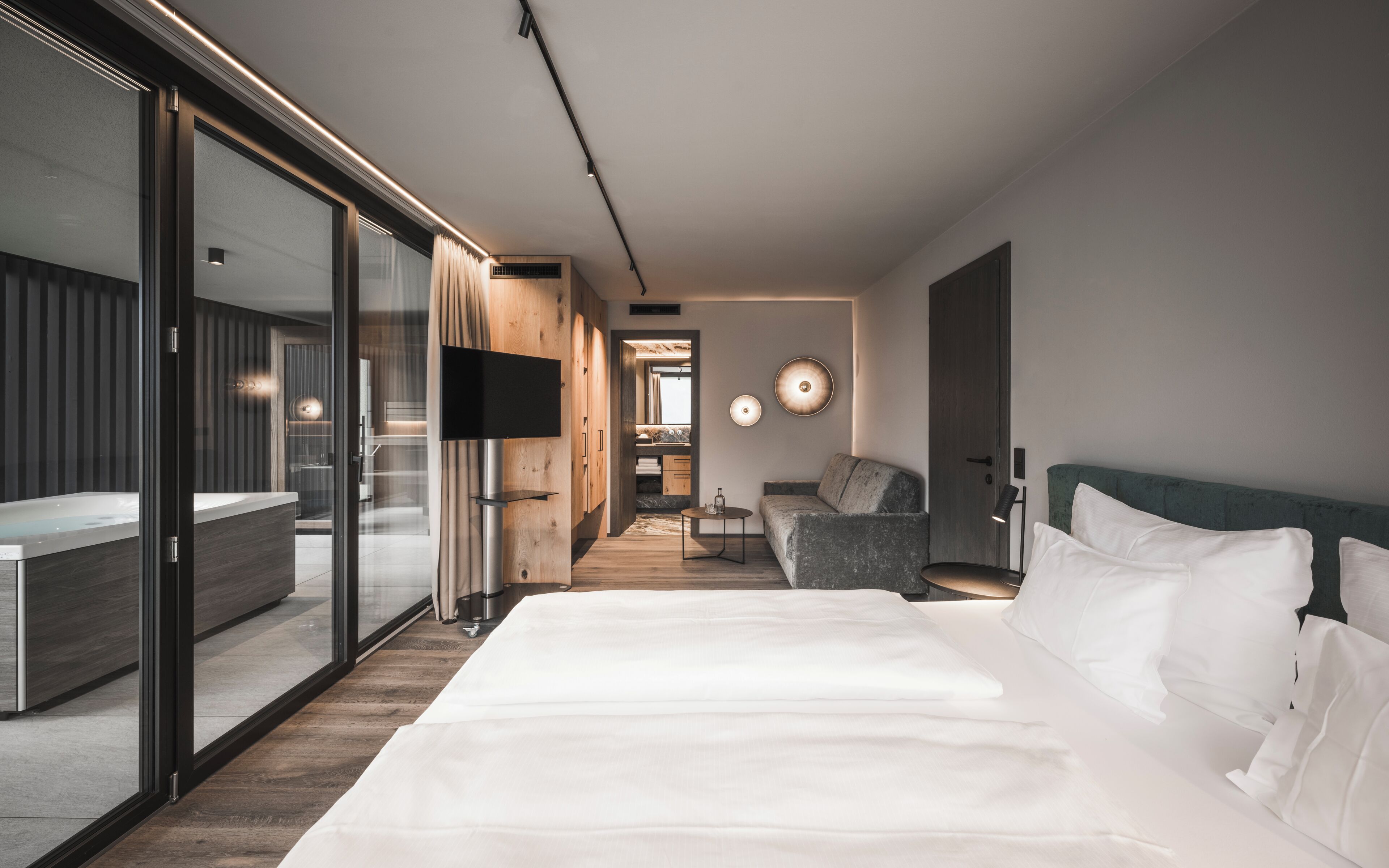 A modern hotel room with a large bed, a seating area with a chair, a television, and a view into the adjacent room.