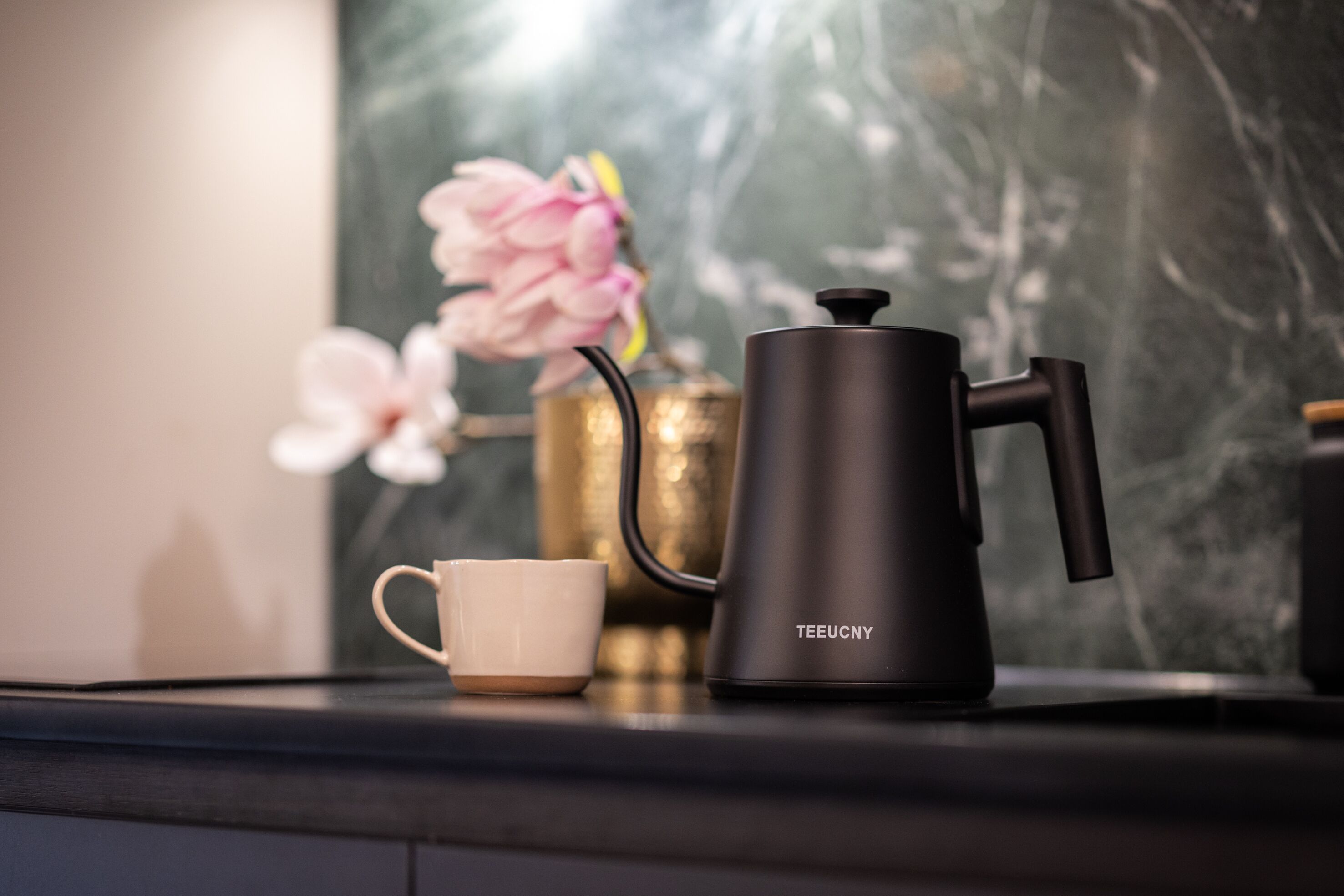 A black coffee maker with a handle and spout, a white cup, and a vase with pink flowers on a dark countertop.