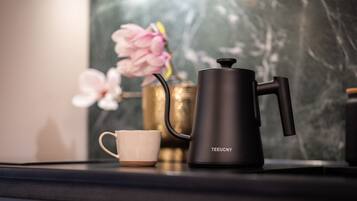 A black coffee maker with a handle and spout, a white cup, and a vase with pink flowers on a dark countertop.