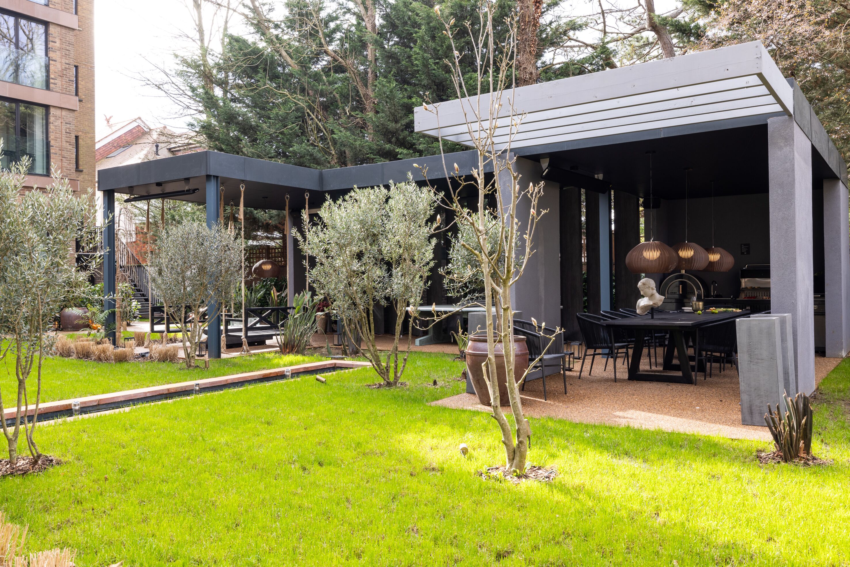 Un área de comedor exterior moderna con porche cubierto, una parrilla y un jardín bien cuidado.