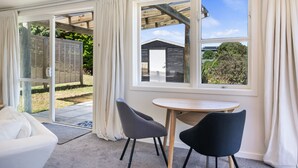 A room with a white sofa, two chairs, a round table, and a view through large windows to the outside. - Hicks Haven - Waikanae Beach Holiday Studio (Waikanae)
