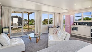 A modern, well-lit living room with a glass coffee table, white leather sofas, and a kitchen with white cabinets and a pink refrigerator. - Hicks Haven - Waikanae Beach Holiday Studio (Waikanae)