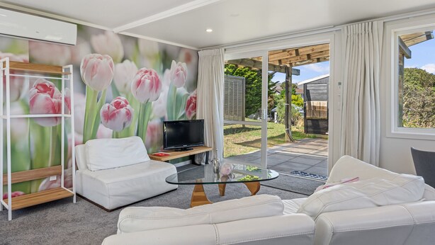 A modern living room with a white sofa, a glass coffee table, and a flat-screen TV. There is a mural of pink tulips on the wall, and a view of the outdoors through large windows. - Hicks Haven - Waikanae Beach Holiday Studio (Waikanae)