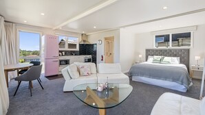 A modern hotel room with a kitchenette, a glass coffee table, a sofa, a bed, and a view of the outdoors. - Hicks Haven - Waikanae Beach Holiday Studio (Waikanae)