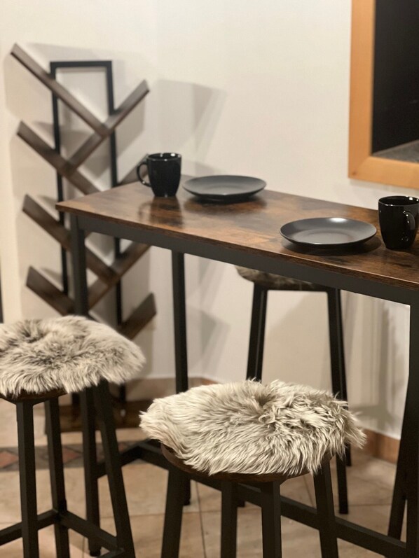 A modern bar area with a wooden table, two stools with fur cushions, and black mugs and plates.