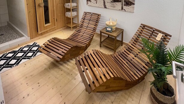 A wooden-floored spa room with two wooden loungers, a small table with a bowl, a towel rack, and a painting of a beach scene on the wall.