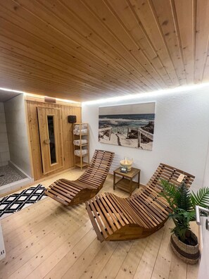 A wooden-floored spa room with two wooden loungers, a small table with a bowl, a towel rack, and a painting of a beach scene on the wall.