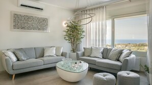 Ein modernes Wohnzimmer mit zwei grauen Sofas, einem Glastisch und einem großen Fenster mit Blick auf das Meer.