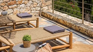 A stone wall with a woven bench and cushions, a round table with a plant, and a view of the sea and mountains. - Villa Eugenia Sunrise (Kefalonia)