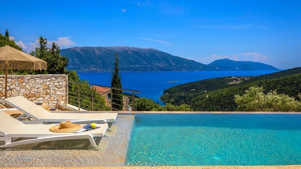 A clear blue swimming pool with lounge chairs and a stone wall, overlooking a lake and mountains. - Villa Eugenia Sunrise (Kefalonia)