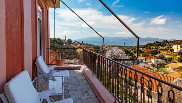 Un balcón con sillas blancas y vista a un pueblo con casas de tejados rojos y montañas al fondo.