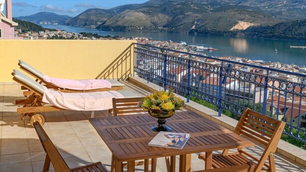 A terrace with a table and chairs, a vase of flowers, and a view of a lake and mountains. - Entheos City Panorama Argostoli (Kefalonia)