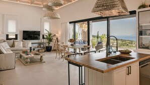 A spacious living area with a large window, a dining table, and a kitchen with a wooden countertop. - Casa Grec Villa Kefalonia (Kefalonia)