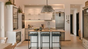 A modern kitchen with white cabinetry, a central island with bar stools, a stainless steel refrigerator, and a hanging pendant light. - Casa Grec Villa Kefalonia (Kefalonia)
