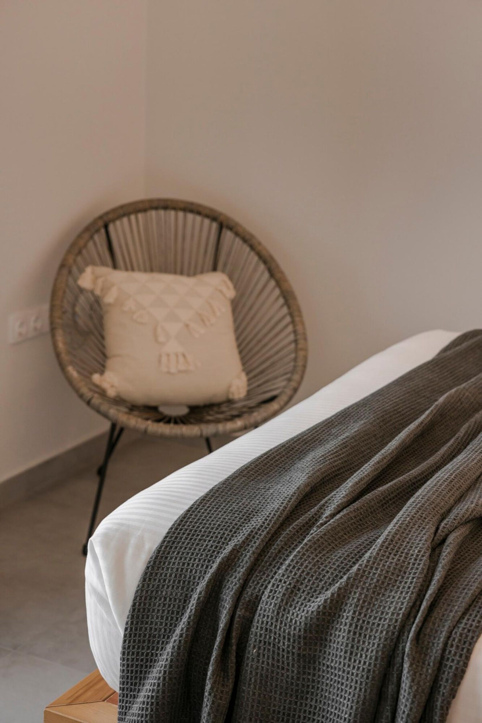 Una cama con sábana blanca y manta gris oscuro, una silla de mimbre con cojín blanco, y una pared lisa con toma de corriente.