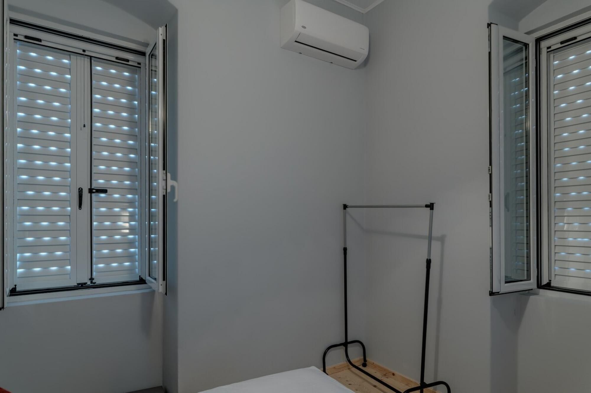 Una habitación con cama, ventana con persianas, equipo de aire acondicionado y un estante metálico.