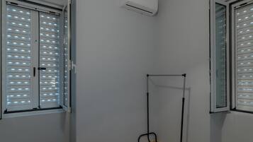 Una habitación con cama, ventana con persianas, equipo de aire acondicionado y un estante metálico.