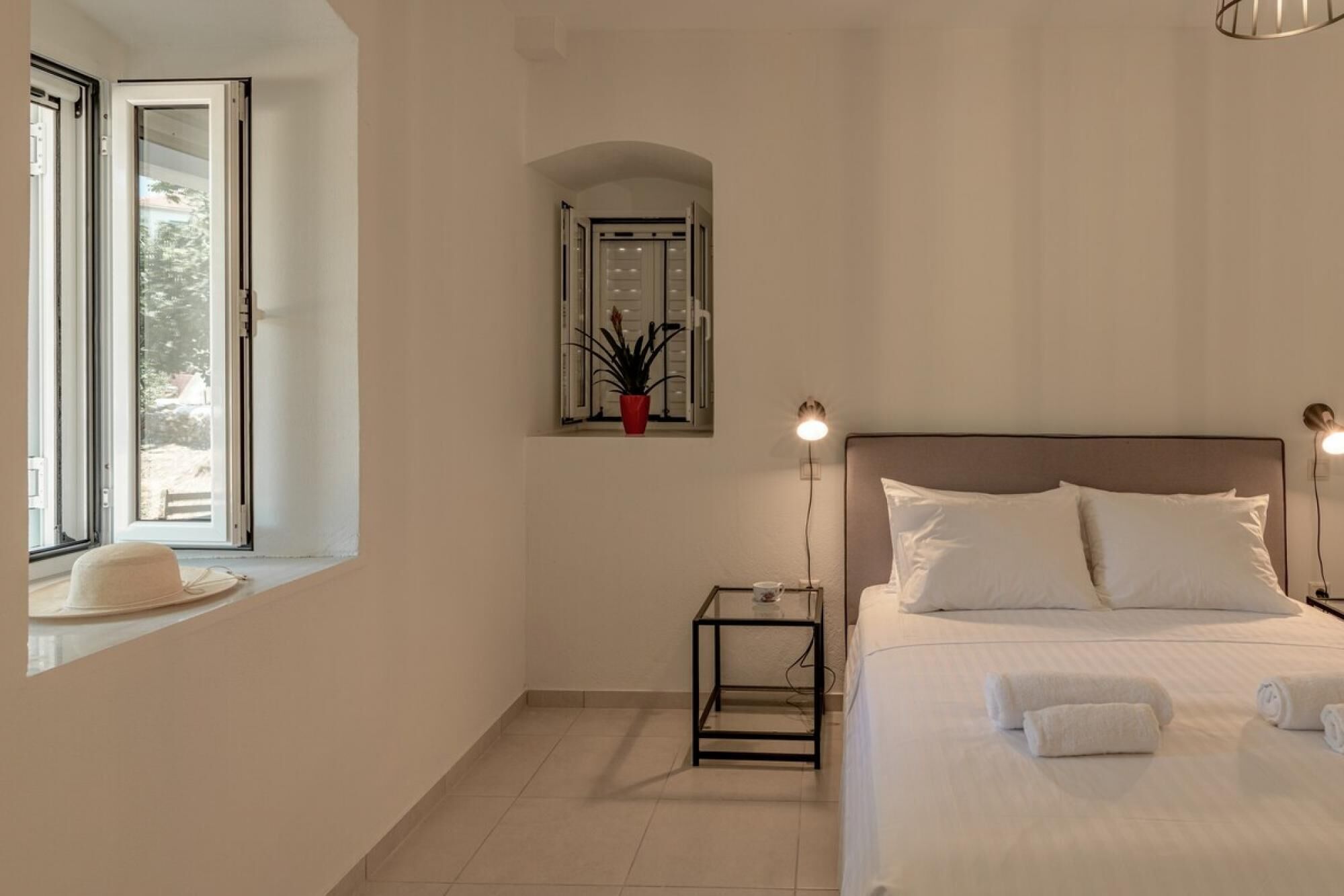 Un dormitorio con cama, lámparas de noche, una ventana y una mesita con una planta.