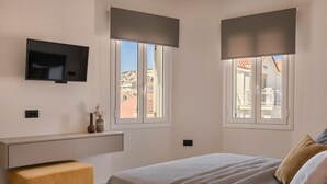 Een slaapkamer met een bed, een aan de muur bevestigde televisie, een nachtkastje en twee ramen met jaloezieën.