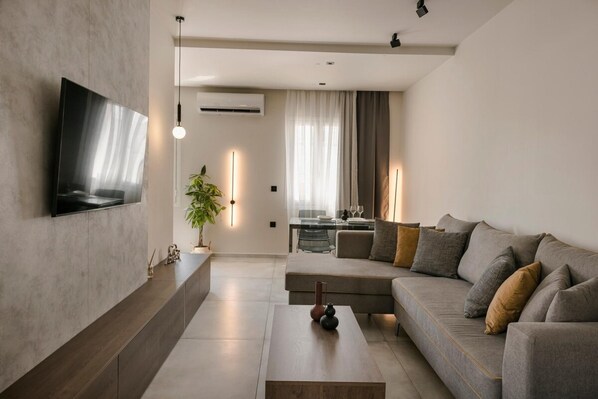 A modern living room with a flat-screen TV mounted on the wall, a sofa, a coffee table, and a dining area in the background. - Argostoli Cityscape Apartment (Kefalonia)