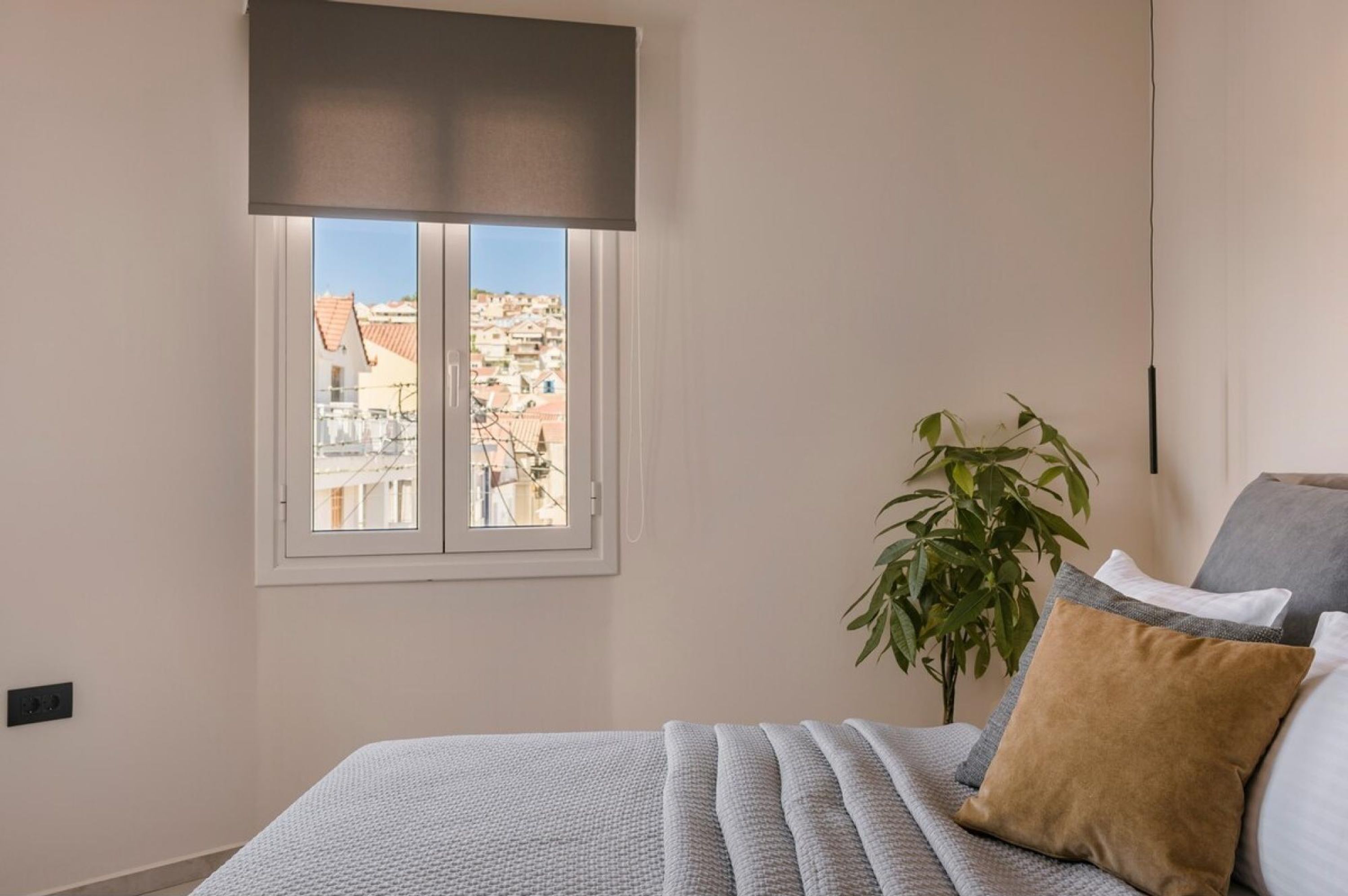 Um quarto com cama, janela com vista para edifícios, uma planta e uma luminária.