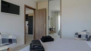 Moderni hotellihuone, jossa on suuri sänky, oleskelualue tuolilla, seinälle asennettu taulutelevisio sekä peili, johon heijastuu kylpyhuone ja keittiönurkkaus.