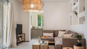 A modern living room with a sofa, coffee table, and a television. There is a kitchen in the background and a large window with curtains.