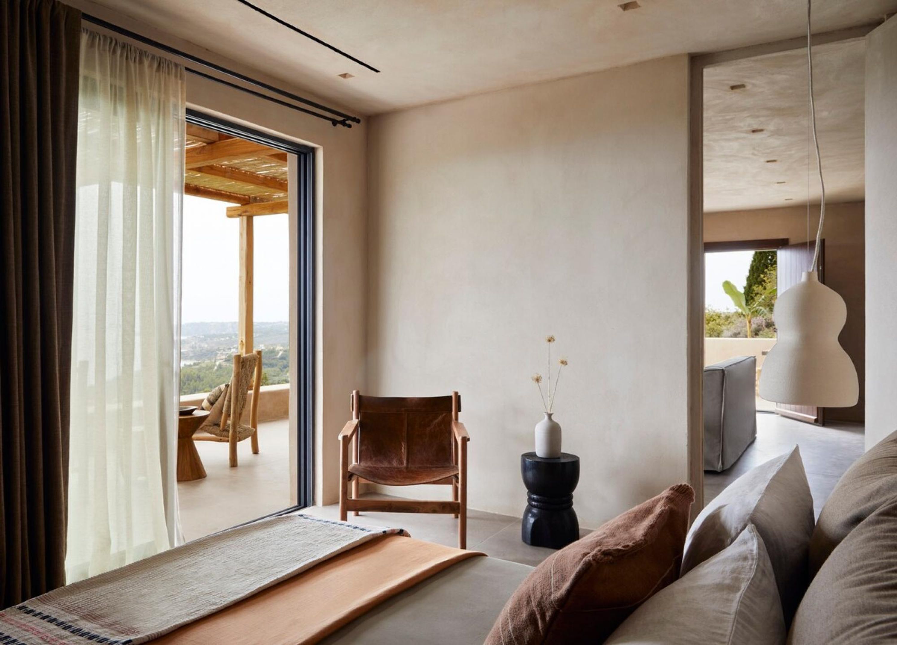 Een moderne slaapkamer met een grote schuifdeur naar een balkon, een comfortabel bed met kussens, een kleine houten stoel en een vaas met bloemen.