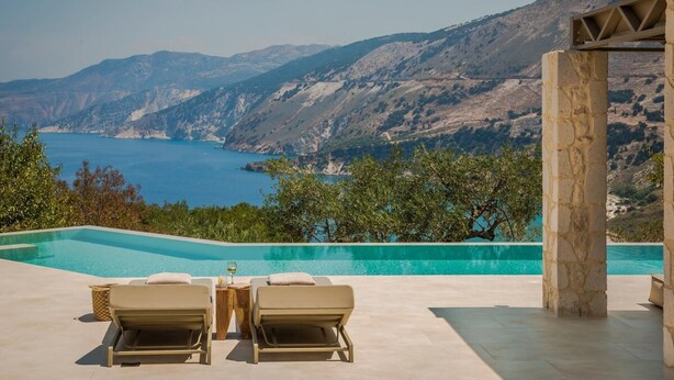 A pool area with two lounge chairs and a table, overlooking a mountainous landscape and a body of water.