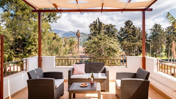 A covered outdoor seating area with wicker furniture and a table, overlooking a mountainous landscape. - Entheos City Center Kefalonia (Kefalonia)