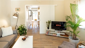A modern living room with a sofa, coffee table, and a TV on a stand. There is a dining area in the background with a table and chairs. - Entheos City Center Kefalonia (Kefalonia)