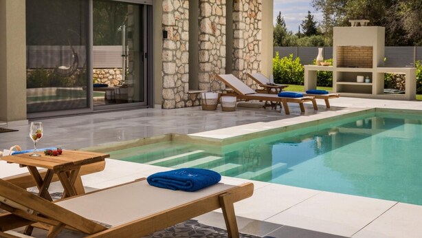 A modern outdoor pool area with lounge chairs, a table with a glass of wine, and a stone wall. - Villa Helena - Ionian Treasure Villas (Kefalonia)
