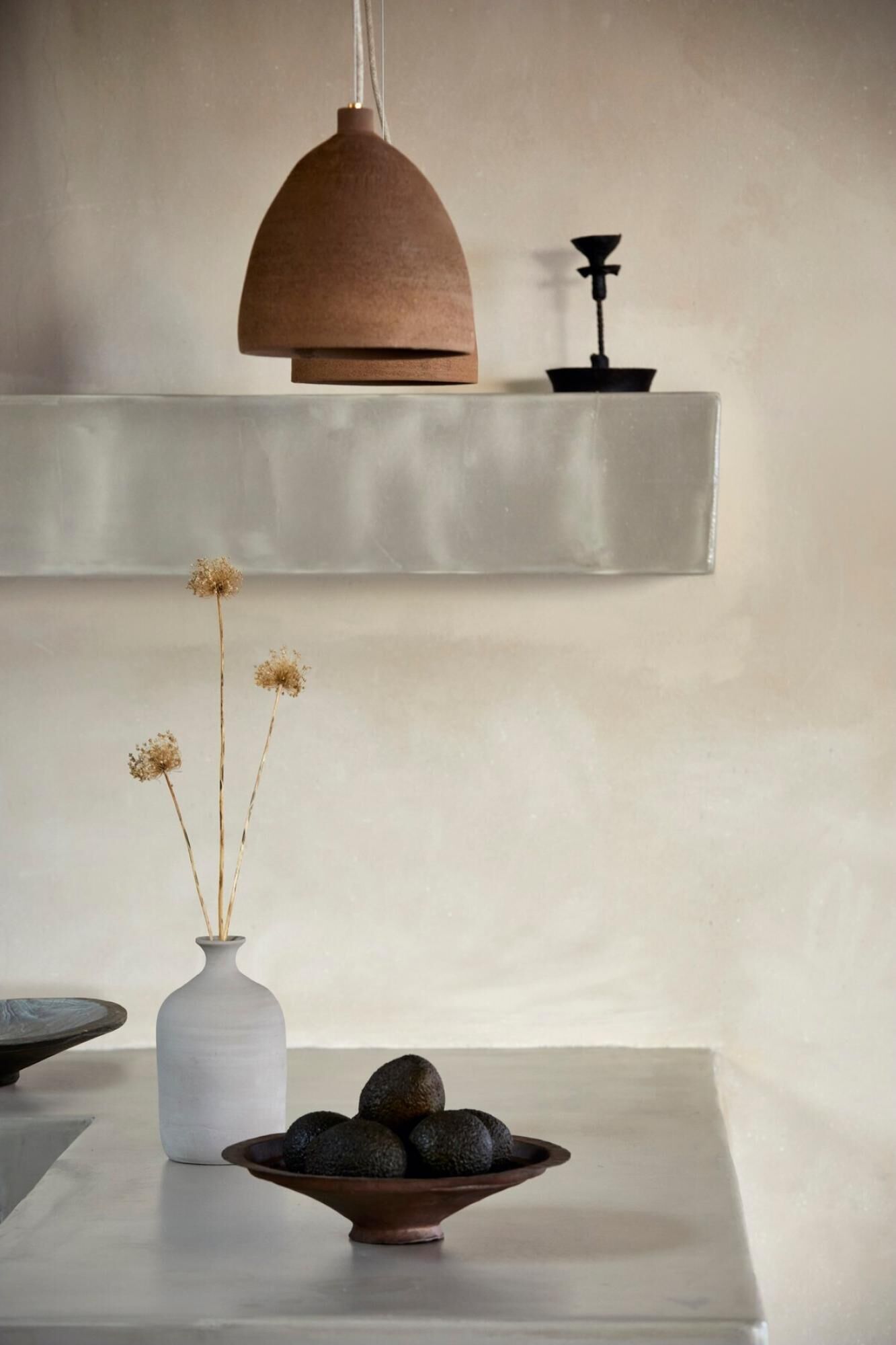 Un intérieur minimaliste avec une table blanche, un vase blanc contenant des fleurs séchées, un bol rempli d’objets sombres et un luminaire suspendu.