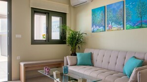 A living room with a beige sofa, a glass coffee table, a bench, and a potted plant. There are three paintings on the wall. - Arktos Sunset Villa (Kefalonia)
