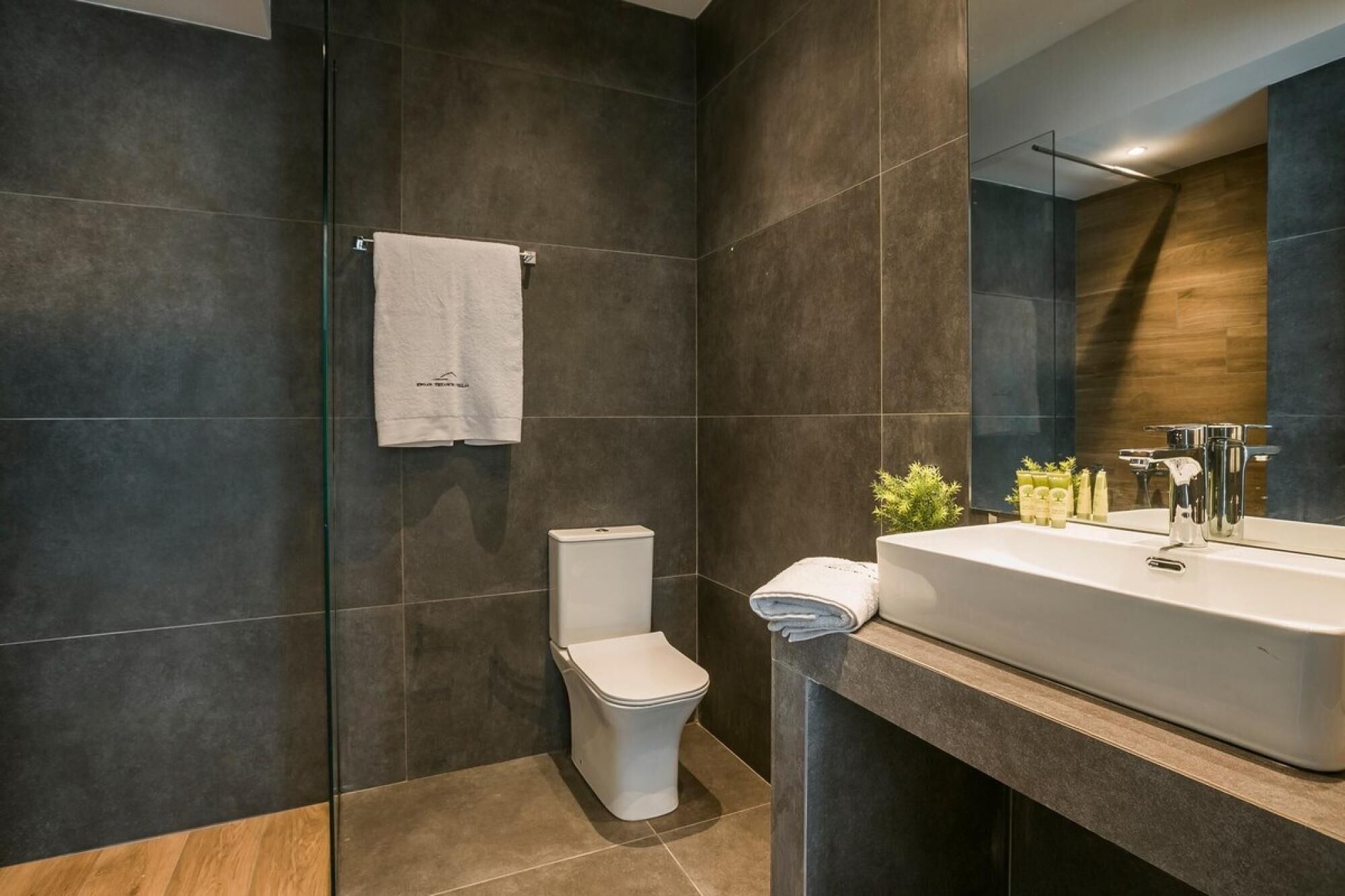 A modern bathroom with dark tiles, a white toilet, a sink with a mirror, and a towel hanging on the wall.